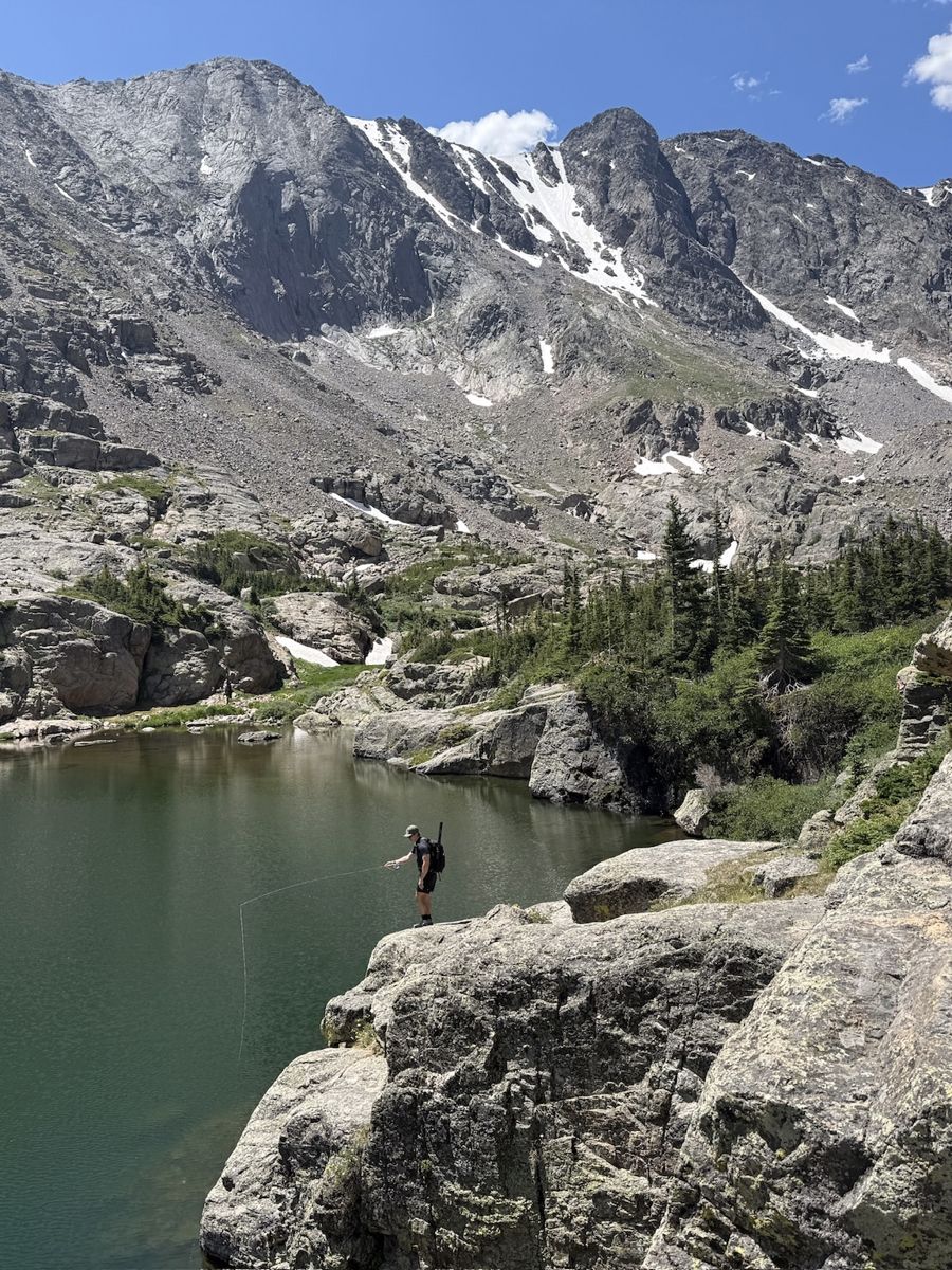Tom casting a fly rod from a rock ledge into an alpine lake below snow-streaked peaks